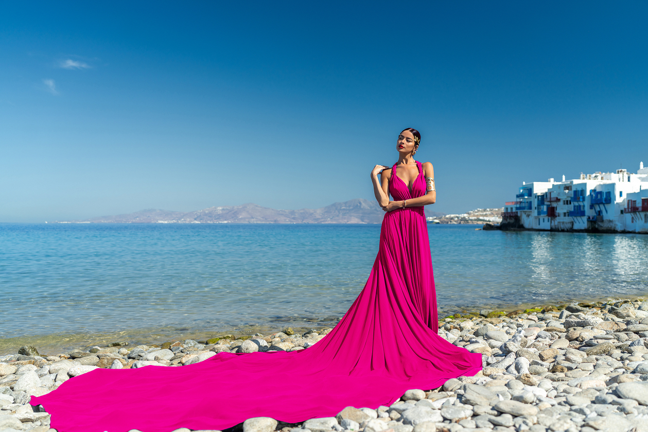 A woman in a flowing fuchsia gown poses elegantly on a rocky beach in Mykonos, with the vibrant colors of Little Venice and the clear blue Aegean Sea providing a stunning backdrop.